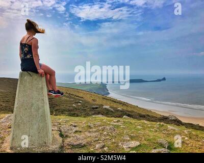 Junge Frau sitzt auf der "Trig point auf Rhossili Downs genießen die Aussicht über Rhossili Bay, Gower, Wales, April. Stockfoto