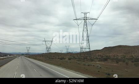 Strommasten und hohe Spannung elektrische Leitungen Kreuzung Freeway und verschwinden in den Bergen, Apple Valley Region, Kalifornien. Bewölkter Tag Stockfoto