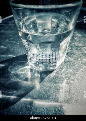 Klar teilweise vollen Glas Wasser auf Metall Terrasse Tisch im hellen Sonnenschein in einem städtischen Umfeld. Stockfoto