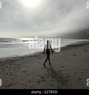 Silhouette eines bärtigen Mannes trug nur Shorts, entlang einem sandigen Britischen Strand bei Ebbe mit dem Abend Sonne glitzert das Meer und glänzend wie ein Feuerball am Himmel Stockfoto