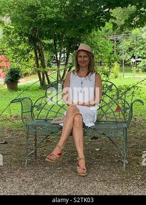 Die Frau trug ein weißes Kleid und einen Strohhut sitzen auf einer Bank in einem Park. Stockfoto