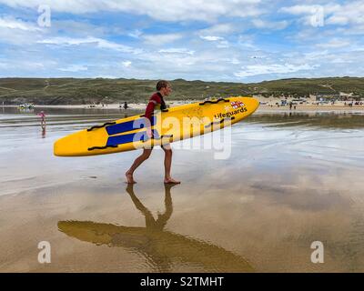 RNLI Rettungsschwimmer zu Fuß auf perran Strand tragen gelbe Surf Board in Cornwall. Stockfoto