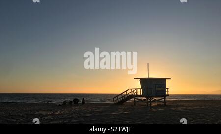 HERMOSA Beach, CA, May 2019: Silhouette Rettungsschwimmer Hütte und die Leute sitzen auf Stühlen neben Pazifischen Ozean bei Sonnenuntergang Stockfoto