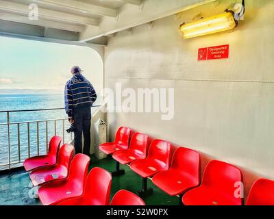 Mann stand allein auf dem Deck der Fähre in der Nähe von Zeilen der leeren Sitze aus rotem Kunststoff, Innere Hebriden, Schottland Stockfoto