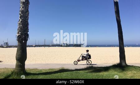 LOS ANGELES, Ca, May 2019: Radfahrer auf Recumbent Bike Fahrten entlang der Promenade zwischen Palmen am Venice Beach. Pier und das Meer im Hintergrund Stockfoto