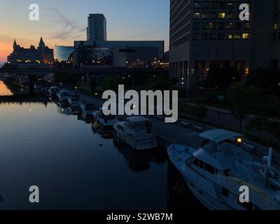 Die Boote in der Rideau Canal, der Innenstadt von Ottawa, der Hauptstadt Kanadas, frühe Nacht, 18. August 2019. Das Chateau Laurier Silhouette ist weit weg gegen den Sonnenuntergang Himmel gesehen. Stockfoto