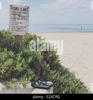 Flip Flops Rest in der Nähe ein Zeichen über die Fischerei auf Pismo Muscheln. Eine Person gesehen, die in das Wasser in der Ferne. Carpinteria State Beach, Carpinteria, Kalifornien, USA Stockfoto