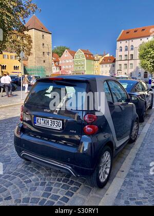 Smart Auto und andere Autos entlang einer Straße mit Kopfsteinpflaster mit bunten Gebäuden und Geschäften in Regensburg, Deutschland geparkt. Stockfoto