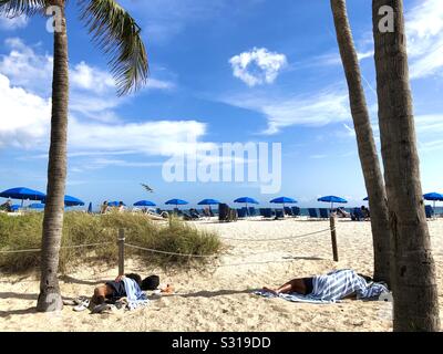 Zwei obdachlose Männer schlafen auf den Sand, während Touristen unter Sonnenschirmen sitzen und auf Liegestühlen entlang des Ocean Beach in Florida. Stockfoto