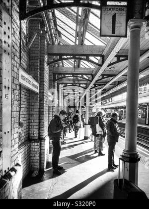 Plattform 1 der Londoner U-Bahnstation High Street Kensington entfernt, von dem aus die Passagiere warten auf den nächsten Zug. Helles Sonnenlicht wirft lange Schatten dieser Szene, in Schwarz und Weiß. Stockfoto