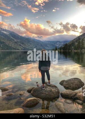 Schöner Blick auf den See, mit einer Dame, die auf einem Felsen steht und den schönen Sonnenuntergang mitnimmt. Stockfoto