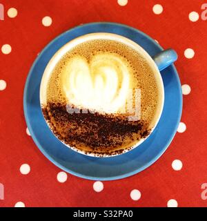 Blick von oben auf einen matten Cappuccino-Kaffee mit Herzform auf der Oberfläche in einer farbenfrohen Tasse und Untertasse mit heißem Getränk und Kopierraum Stockfoto