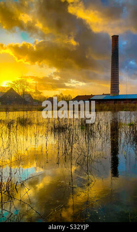 Dramatischer und bunter Sonnenuntergang und Ruinen einer alten Steinfabrik spiegelten sich im Wasser einer Pfütze im Gras wider Stockfoto