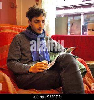 Junger Mann liest eine Zeitschrift, während er in einem Café-Sessel sitzt. Stockfoto