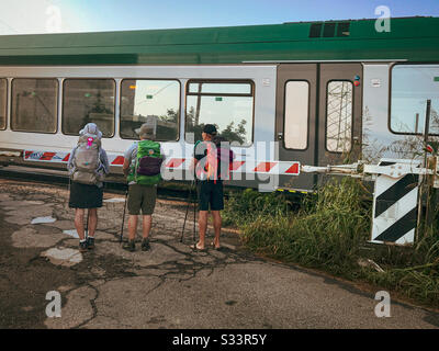 Drei Rucksacktouristen, eine Frau und zwei Männer, die an einem Bahnübergang warten, bis ein Zug vorbei fährt. Stockfoto