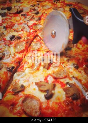 Frisch gebackene Pizzakuchen mit Wurst- und Pilzbesatz, die von einem Pizza-Schneiderad in Scheiben geschnitten werden Stockfoto