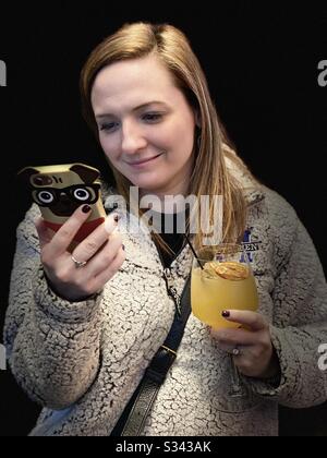 Isolierte kaukasische junge Frau vor schwarzem Hintergrund, die ihr Handy in der einen Hand betrachtet und ein alkoholisches Getränk in der anderen hält, während sie lächelt. Stockfoto
