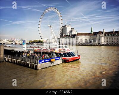 Am Westminster Pier auf der Themse ist ein Stadtkreuzfahrtschiff zu sehen, mit Blick auf das Millennium Wheel und die Flugzeugkontrails in London Stockfoto