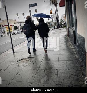 LOS ANGELES, CA, MAR 2020: Junges Paar mit Mann, das Regenschirm über seiner Freundin trägt, während sie durch nasse Straßen in Korea-Stadt laufen Stockfoto