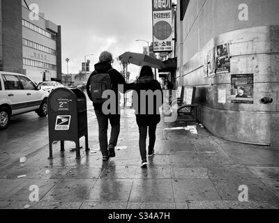 LOS ANGELES, CA, MAR 2020: Junges Paar mit Mann, das Regenschirm über seiner Freundin trägt, während sie durch nasse Straßen in Korea-Stadt laufen Stockfoto