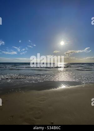 Porträtansicht der niedrigen Sonne über dem Pazifischen Ozean mit spärlichen Wolken am Himmel, sanfte Wellen, die den Sandstrand im Vordergrund lappen Stockfoto