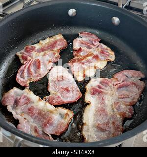 Ein Blick auf den Speck, der in der Bratpfanne auf einem Kochfeld sizzt. Speckscheiben zum Frühstück braten. Stockfoto