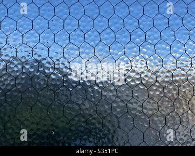 Gestelztes, opakes Sicherheitsglas mit durchgehendes Drahtgitter, undeutlicher blauer Himmel und Stadt, die durch ihn kaum sichtbar sind, wodurch eine abstrakte Hintergrundtextur entsteht. Stockfoto