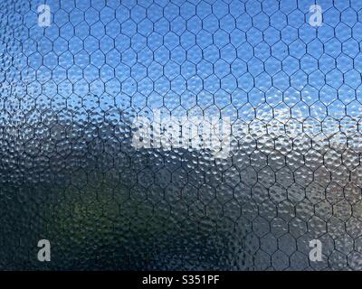 Breit gestelztes, opakes Sicherheitsglas mit durchgehendes Drahtgitter, undeutlicher blauer Himmel und Stadt, die kaum durch ihn sichtbar sind, wodurch eine abstrakte Hintergrundtextur entsteht. Stockfoto