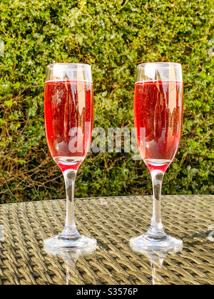 Zwei Flötengläser aus rosa Champagner auf der Glasplatte eines Tisches in einem Garten an einem Sommerabend Stockfoto
