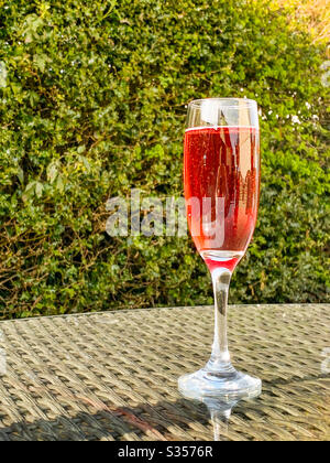 Ein Glas mit einer Flöte aus rosa Champagner auf der Glasplatte eines Tisches in einem Garten an einem Sommerabend Stockfoto
