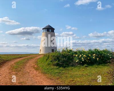 Restauriertes altes Windmühlengebäude auf einem grünen Hügel mit Narzissen und einem gewundenen schlammigen Pfad. Blauer Himmel mit Wolken. Stockfoto