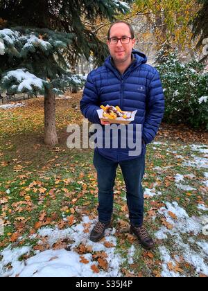 Mann hielt ein Tablett mit frisch zubereiteten Churros, die mit Schokoladensauce bedeckt waren, während er in einem verschneiten Park stand, der von Bäumen umgeben war. Stockfoto