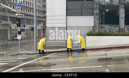 LOS ANGELES, CA, APR 2020: Drei Personen Reinigungscrew in gelben Regenmänteln, die Dachrinnen in der 4th St, Downtown, neben einem Parkhaus, im Regen räumen Stockfoto