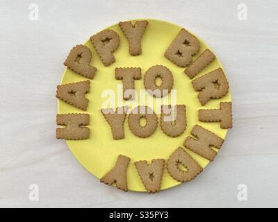 Happy Birthday, kreative Nachricht mit Keksbuchstaben auf einem gelben Teller über einem weißen Holzbrett komponiert Stockfoto