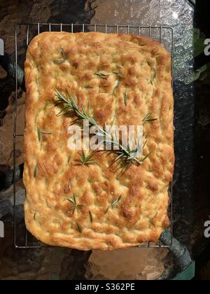 Von oben ein frisch gebackenes, hausgemachtes Focaccia mit Rosmarin auf einem Kuchenregal und einem Tisch mit Glasplatte. Während COVID-Sperrung Stockfoto