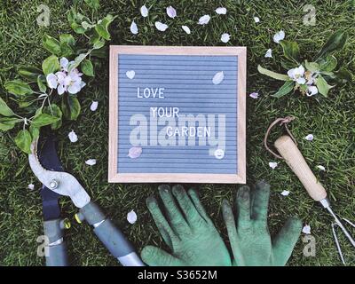 Liebe deinen Garten. Gartenkonzept mit Filzbriefbrett, Handpflaume, schmutzigen Handschuhen und einem beschnittenem Ast mit Blüten von einem Apfelbaum auf einem grünen Rasen Stockfoto