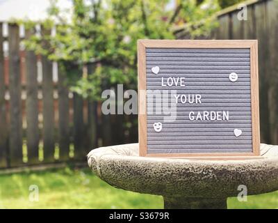 Liebe deinen Garten. Gartenkonzept mit Filztafel auf einem Vogelbad inmitten eines Vorstadtgartens mit Zwergapfelbaum im Hintergrund und Kopierraum. Stockfoto