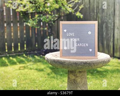 Liebe deinen Garten. Gartenkonzept mit Filztafel auf einem Vogelbad inmitten eines Vorstadtgartens mit Zwergapfelbaum im Hintergrund und Kopierraum. Stockfoto