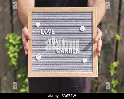 Liebe deinen Garten. Gartenbau-Konzept mit Filz Message Board in den Händen eines kaukasischen Mann steht gegen einen verwitterten Zaun mit überwucherten Efeu im Hintergrund gesehen verschwommen. Stockfoto