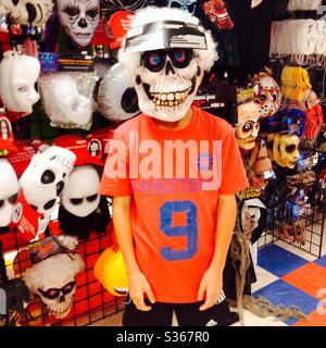 Junger Junge in einem Spielzeugladen versucht auf einer gruseligen Skelett Halloween Maske durch eine große Anzeige von Masken. Stockfoto