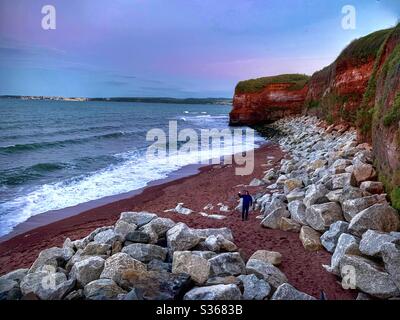 Mann steht an einem Strand bei Flut, umgeben von roten Klippen, Sand, große Felsbrocken und einem stürmischen Meer. Stockfoto