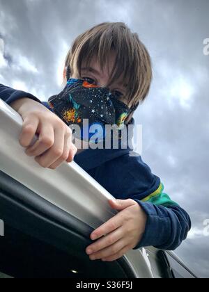 Ein 10-jähriger Junge in einer Maske spielt auf dem Fahrzeug seiner Mutter, während er auf einen Besuch der Zufahrt mit seiner Großmutter während der COVID-19-Quarantäne wartet. Stockfoto