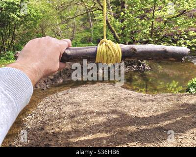 Nahaufnahme von Details eines Mannes, der an einer Seilschaukel hält, die an den Zweigen eines reifen Baumes in einem Wald hängt. Ein kleiner Stab wird mit Knoten als Bügel gebunden, um über Fluß unten zu schwingen Stockfoto