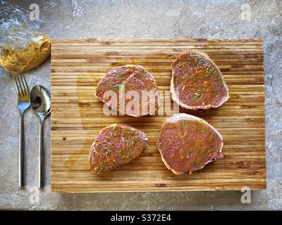 Nahaufnahme von rohen Rindssteaks, mariniert mit Knoblauch, Olivenöl und Gewürzen. Zubereitung von Zutaten für das Abendessen in der Küche auf einer rustikalen Holztheke Stockfoto