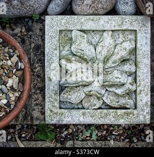 Dekorative Gartenfliesen mit Pflanzendesign neben einem Terrakotta-Topf auf einer niedrigen Ziegelwand. Grüne Färbung der Fliese durch verblasste Orange des Topfes versetzt. Viele Texturen und Farben in Steinen, Ziegel und Kies. Stockfoto