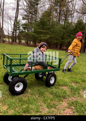 Glückliche ältere Schwester zieht ihren lächelnden kleinen Bruder in einem Wagen in warmen Jacken und Hüten gekleidet. Stockfoto