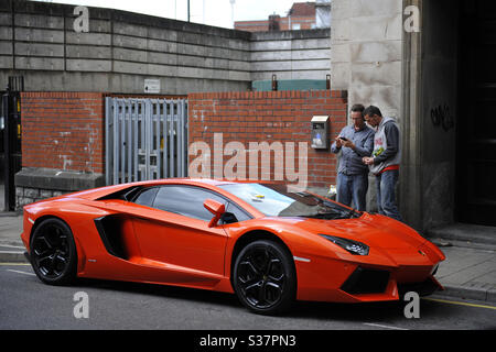 Zwei Männer schauen auf einen orangefarbenen Lamborghini Aventador, der im Zentrum von Cardiff, Wales, Großbritannien, geparkt ist und dort ein Parkschein erhalten hat. Stockfoto