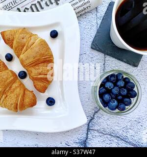 Nahaufnahme von frisch gebackenen Croissants auf einem weißen Teller mit Heidelbeeren und Trauben. Leckeres Frühstück mit Morgenzeitung auf einem Tisch aus Naturstein. Serviert mit Kaffee. Stockfoto