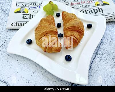 Nahaufnahme von frisch gebackenen Croissants auf einem weißen Teller mit Heidelbeeren und Trauben. Leckeres Frühstück mit Morgenzeitung auf einem Tisch aus Naturstein. Stockfoto