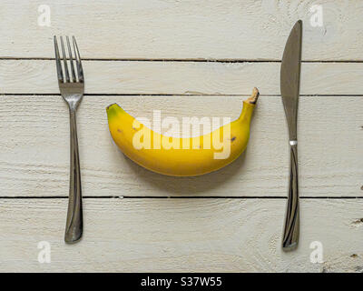 Gelbe Banane mit Messer und Gabel auf weißen Holzbohlen
